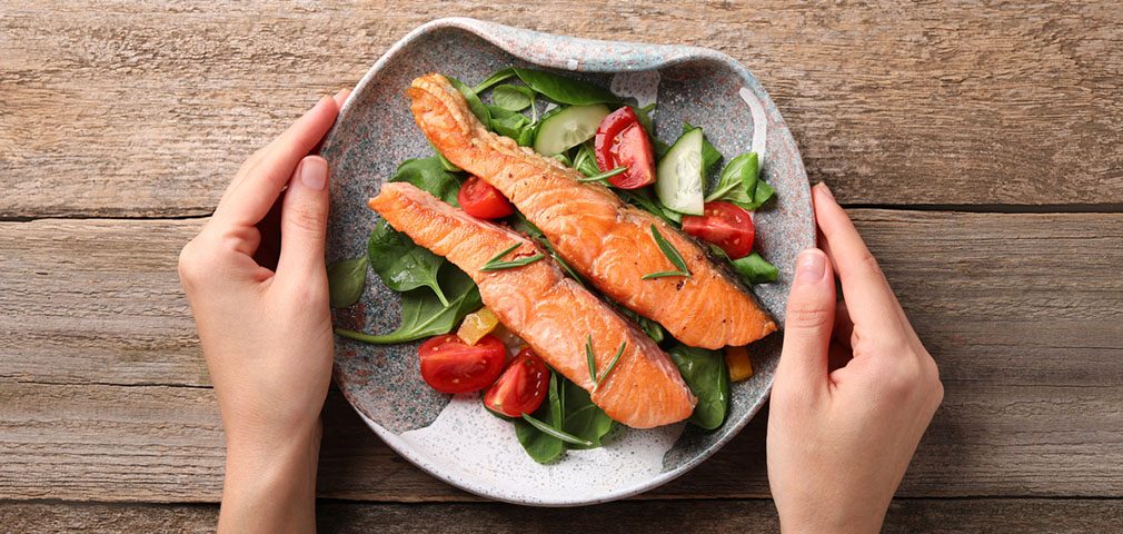 Healthy meal. Woman with plate of tasty grilled salmon, spinach and vegetables at wooden table