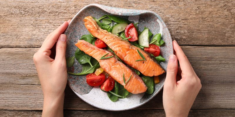 Healthy meal. Woman with plate of tasty grilled salmon, spinach and vegetables at wooden table